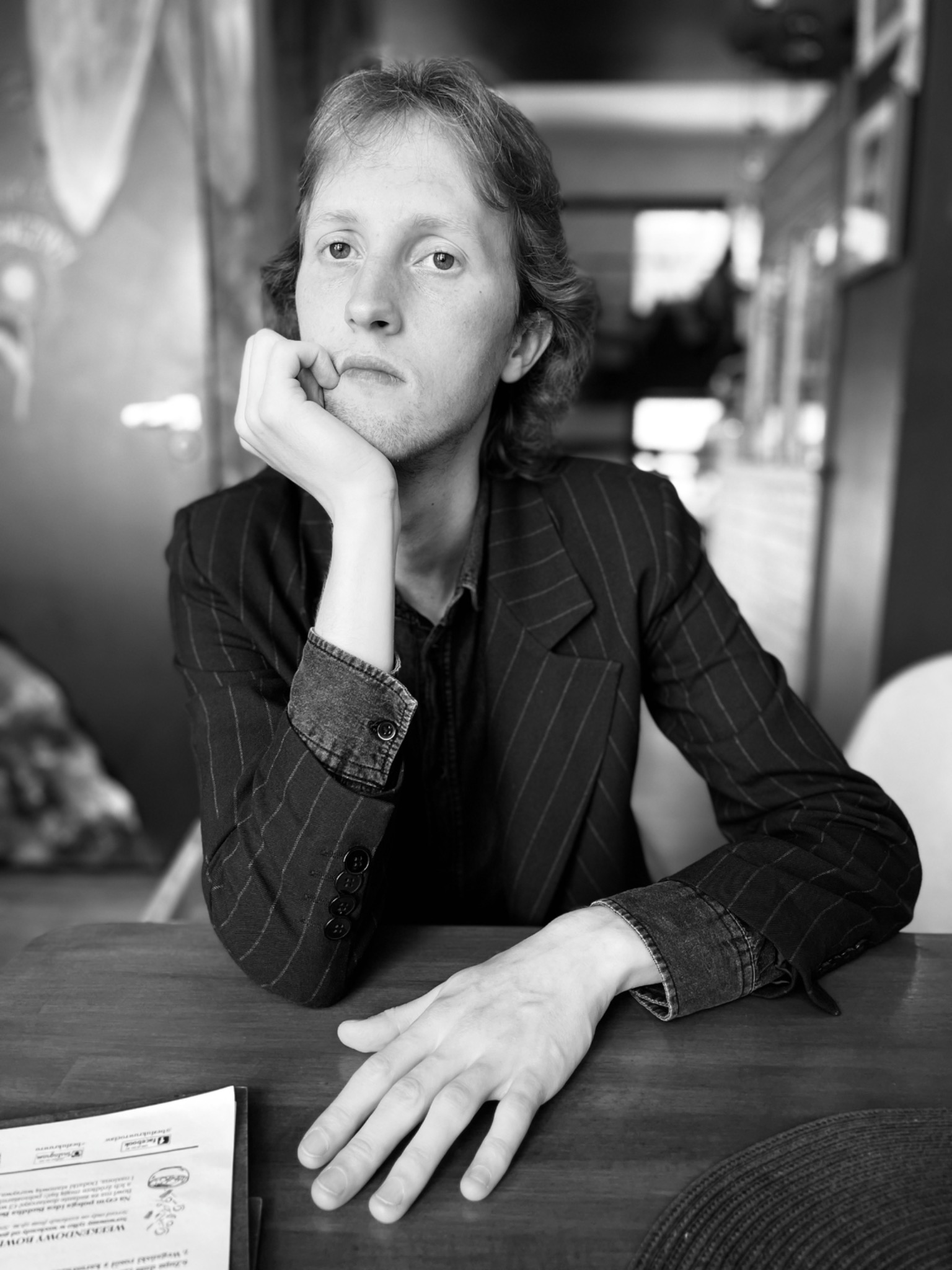 Portret czarno-biały mężczyzny w marynarce w paski, siedzącego przy drewnianym stole z dokumentami, zamyślony wyraz twarzy, oświetlenie górne.