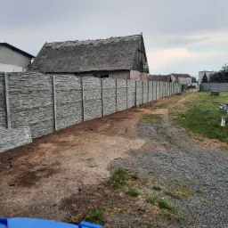 Długi, szary, betonowy płot panelowy z fakturą kamienia, biegnący wzdłuż drogi gruntowej, z budynkiem krytym strzechą w tle.