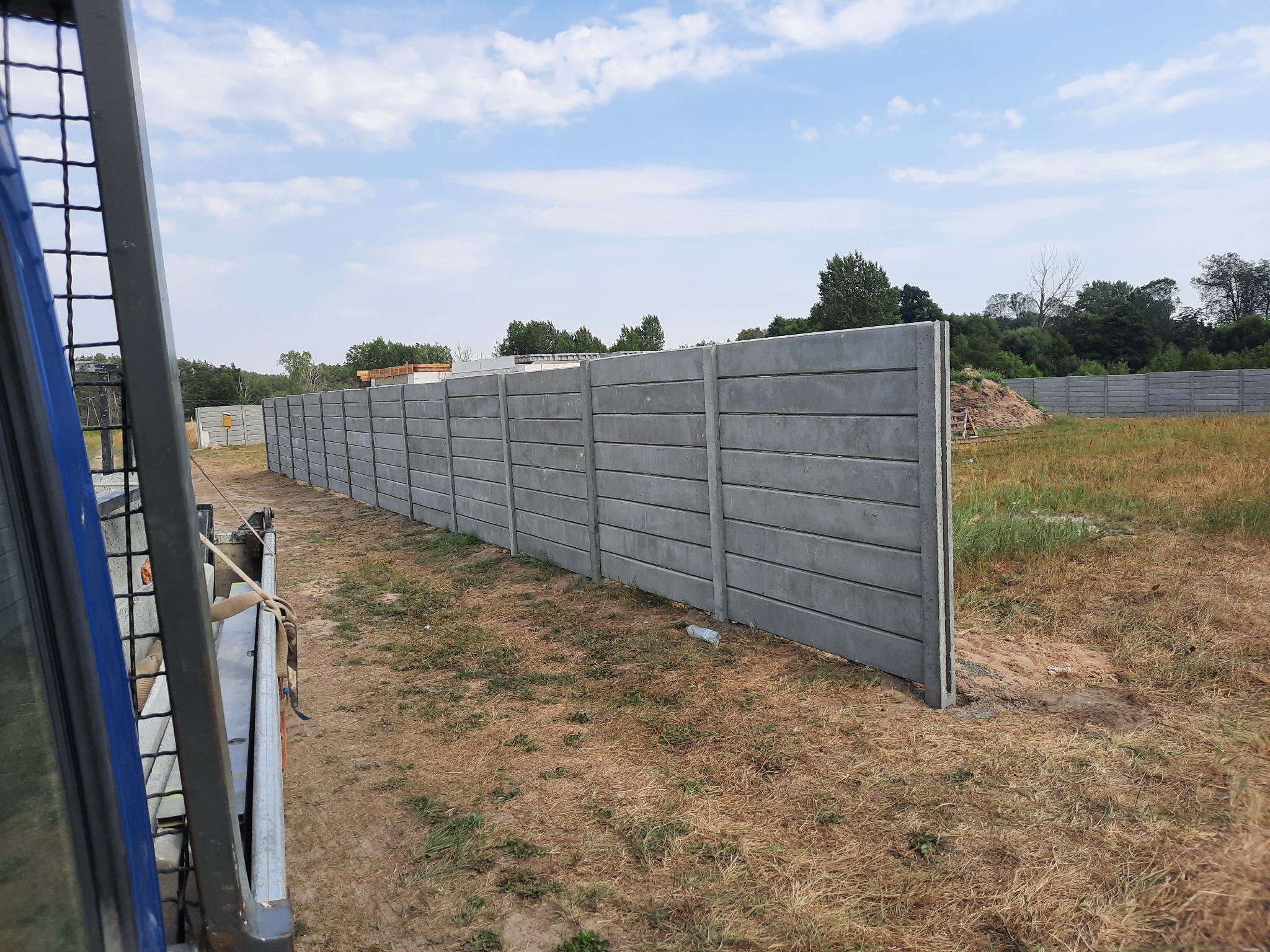 Długa, betonowa, prefabrykowana konstrukcja ogrodzeniowa, widziana z perspektywy pojazdu, z polami i drzewami w tle.