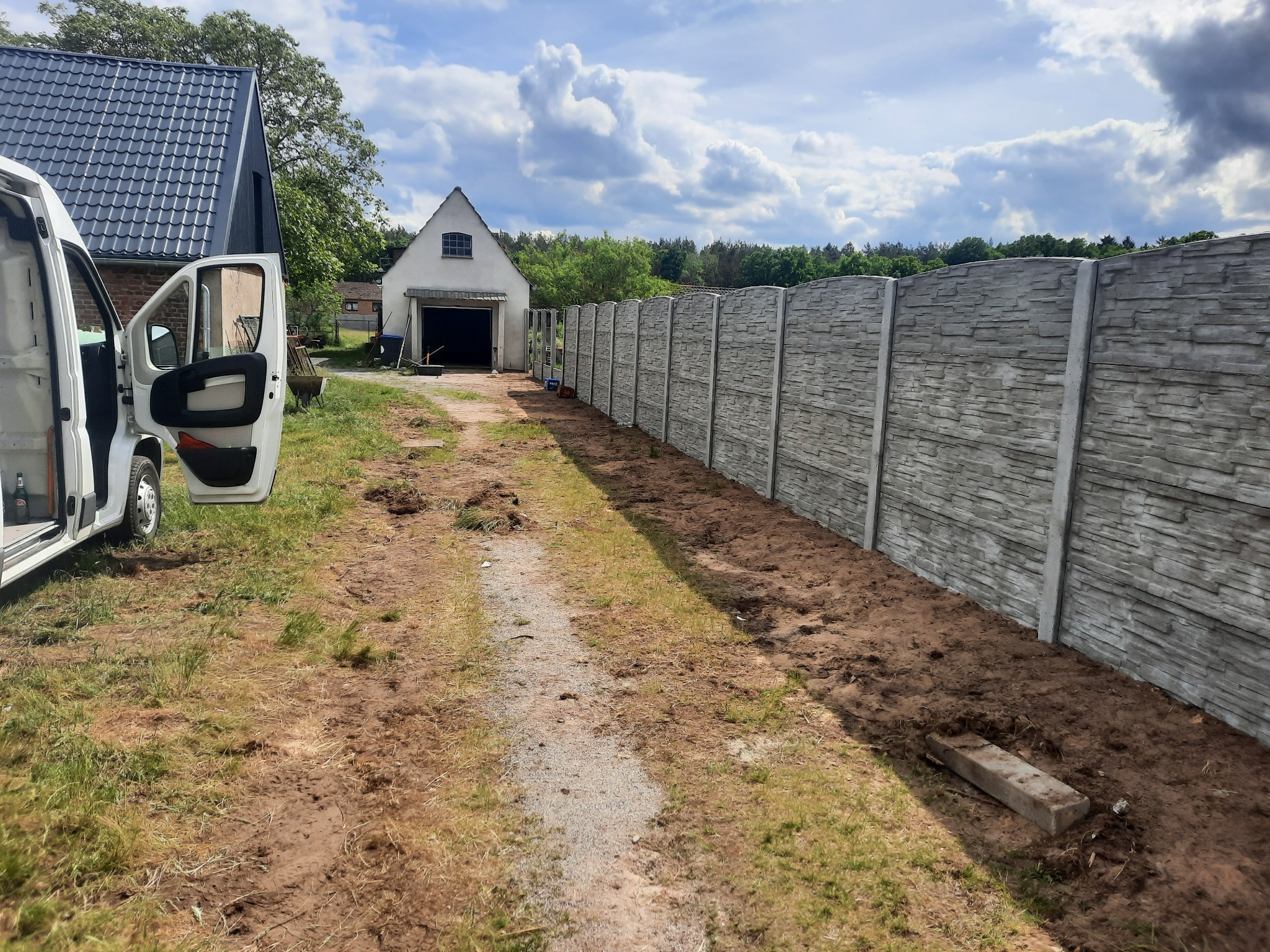 Nowo postawione, betonowe ogrodzenie panelowe z fakturą kamienia, widoczne z perspektywy drogi dojazdowej, z otwartymi drzwiami białego samochodu dostawczego w tle.