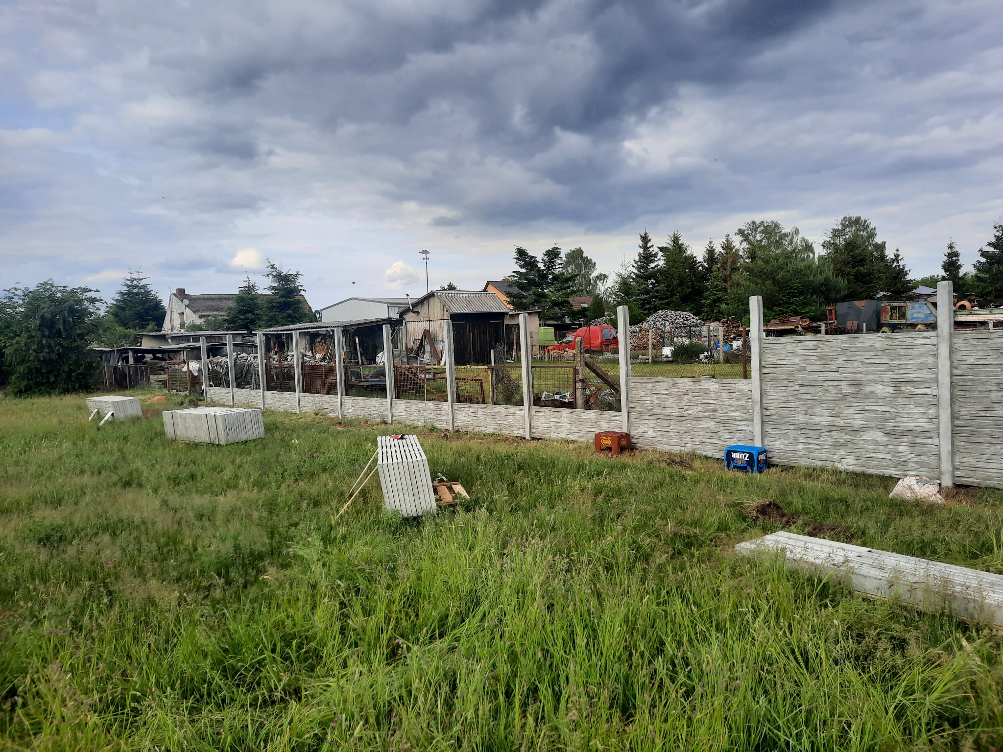 Montaż ogrodzenia panelowego z betonową podmurówką na działce z wysoką trawą i pochmurnym niebem w tle.