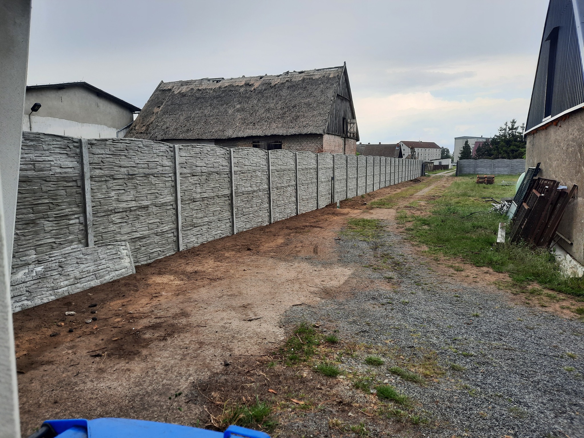 Długi, szary, betonowy płot panelowy z fakturą kamienia, biegnący wzdłuż drogi gruntowej, z budynkiem krytym strzechą w tle.