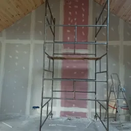 Metalowe rusztowanie na kółkach w pokoju na poddaszu ze skośnym drewnianym sufitem, ściany pokryte płytami gipsowo-kartonowymi, widoczne ślady szpachlowania.