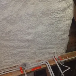 Poddasze ocieplone białą pianką PUR, widoczne rury instalacji elektrycznej i hydraulicznej, zabezpieczone folią i taśmą.