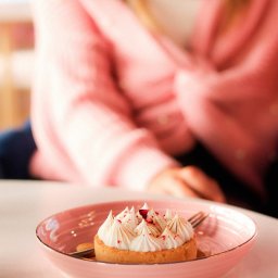 Justyna Skukowska - Kremowa babeczka z przypalonymi końcówkami i posypką z czerwonych płatków na różowym talerzu, w tle osoba w różowej bluzce
