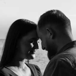 Justyna Skukowska - Czarno-białe zbliżenie profilu kobiety i mężczyzny, dotykających się czołami na tle wody i jasnego nieba, delikatny uśmiech kobiety, broda mężczyzny.