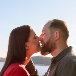 Justyna Skukowska - Profil całującej się pary na tle jasnego nieba i lekko zamglonego krajobrazu z wodą w tle. Kobieta w czerwonej bluzce, mężczyzna w ciemnej koszuli.