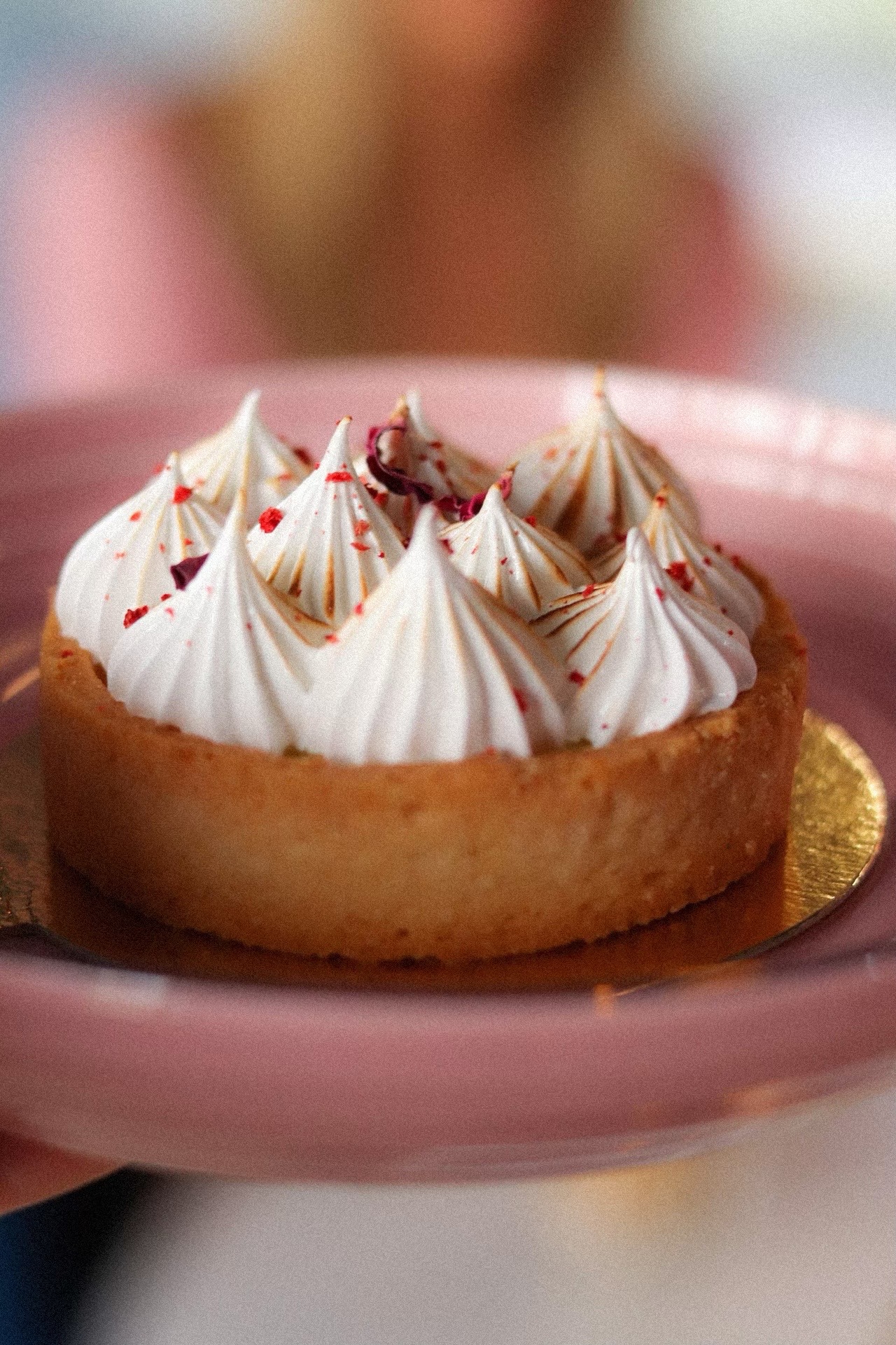 Mini tarta cytrynowa z przypieczoną bezą, posypana czerwonymi płatkami, na różowym talerzyku i złotym podkładzie.