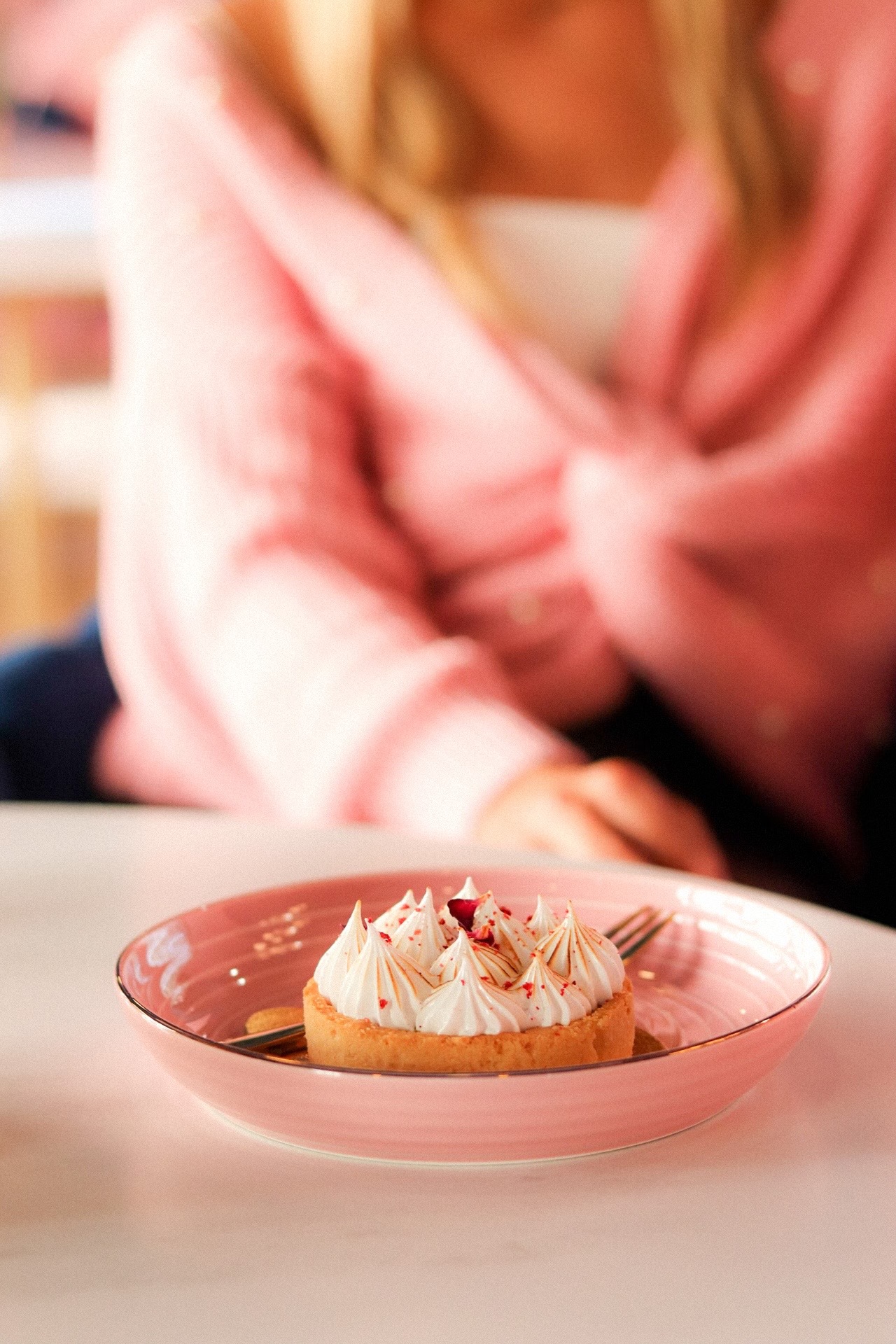 Kremowa babeczka z przypalonymi końcówkami i posypką z czerwonych płatków na różowym talerzu, w tle osoba w różowej bluzce