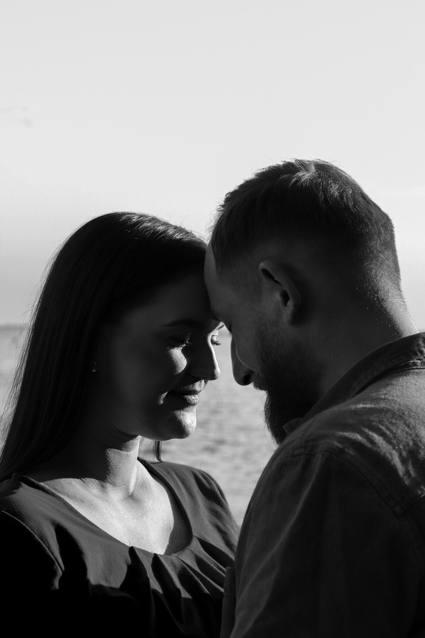 Czarno-białe zbliżenie profilu kobiety i mężczyzny, dotykających się czołami na tle wody i jasnego nieba, delikatny uśmiech kobiety, broda mężczyzny.