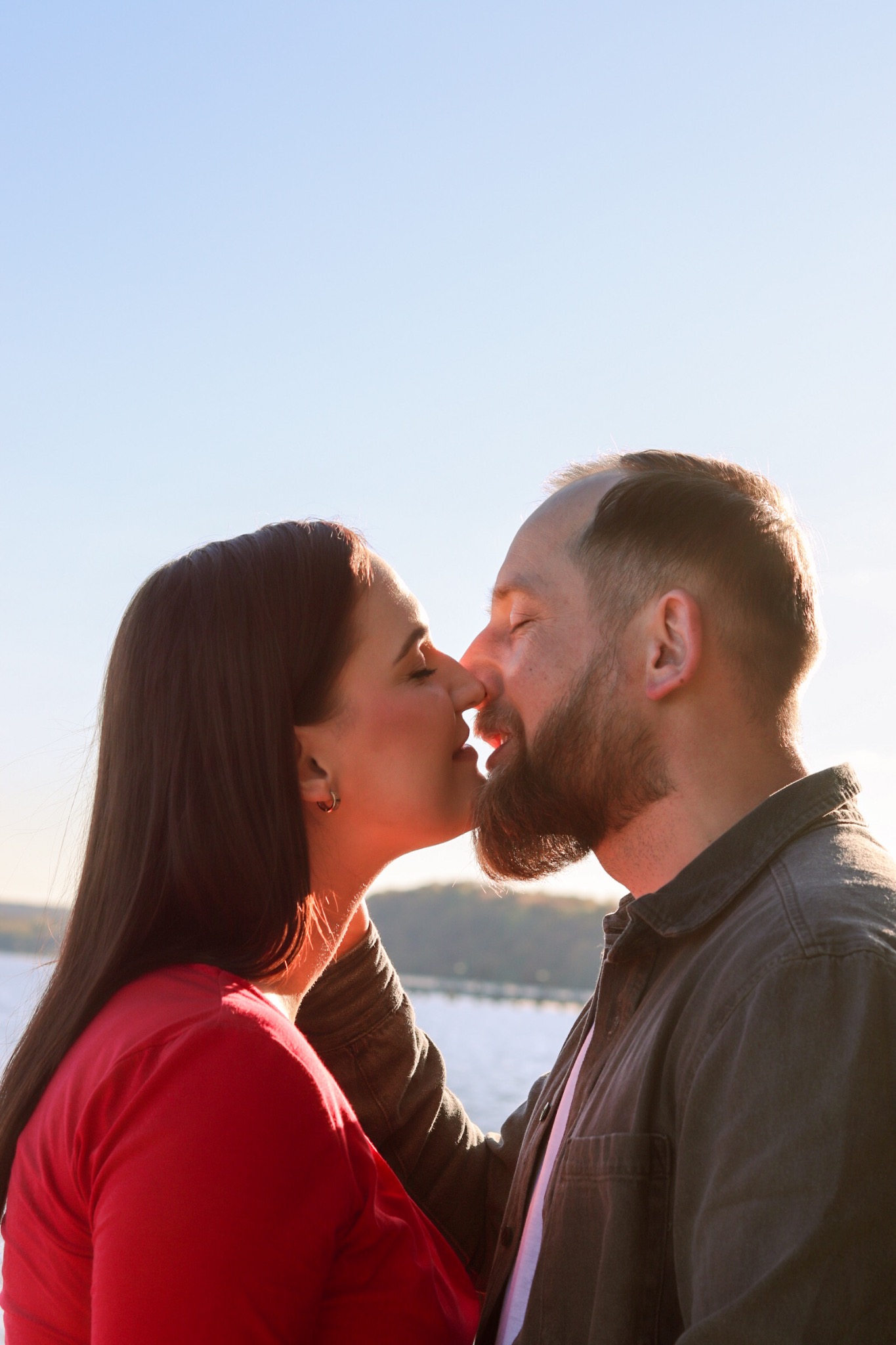 Profil całującej się pary na tle jasnego nieba i lekko zamglonego krajobrazu z wodą w tle. Kobieta w czerwonej bluzce, mężczyzna w ciemnej koszuli.