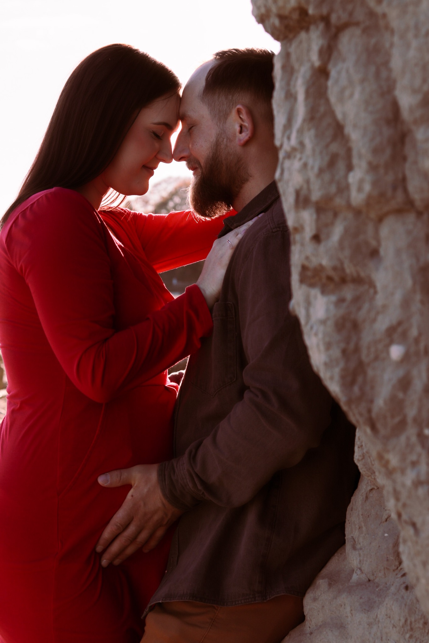Ciężarna kobieta w czerwonej sukience i mężczyzna z brodą opierają się czołami o siebie przy skalnej ścianie w ciepłym świetle zachodzącego słońca.