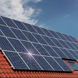 Panele słoneczne na czerwonym dachu domu jednorodzinnego z oknem dachowym, błękitne niebo w tle.