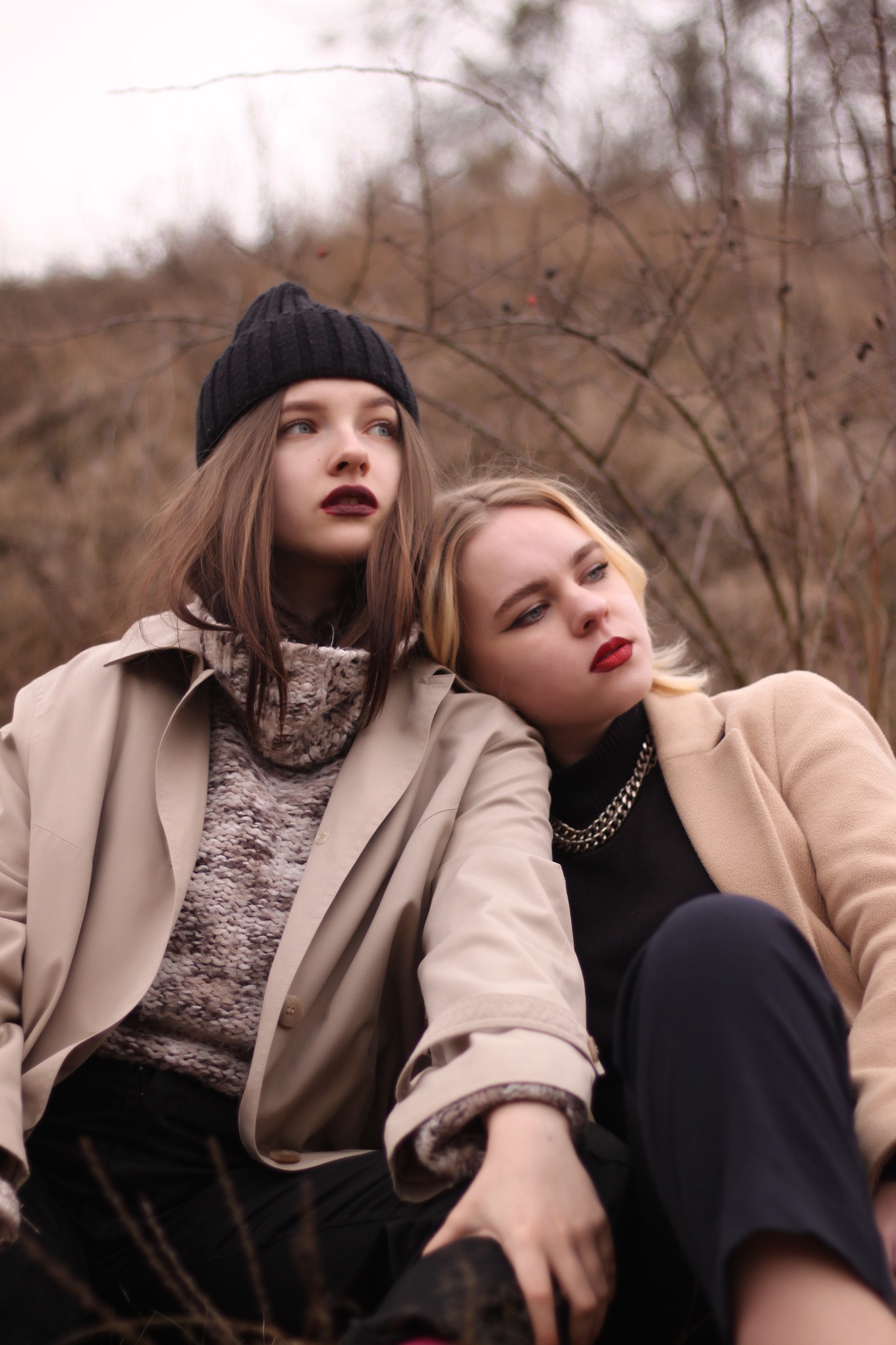 Dwie młode kobiety w jesiennych stylizacjach, beżowych płaszczach i ciemnych ubraniach, siedzą blisko siebie na tle suchych krzewów, patrząc w dal z melancholijnym wyrazem twarzy.