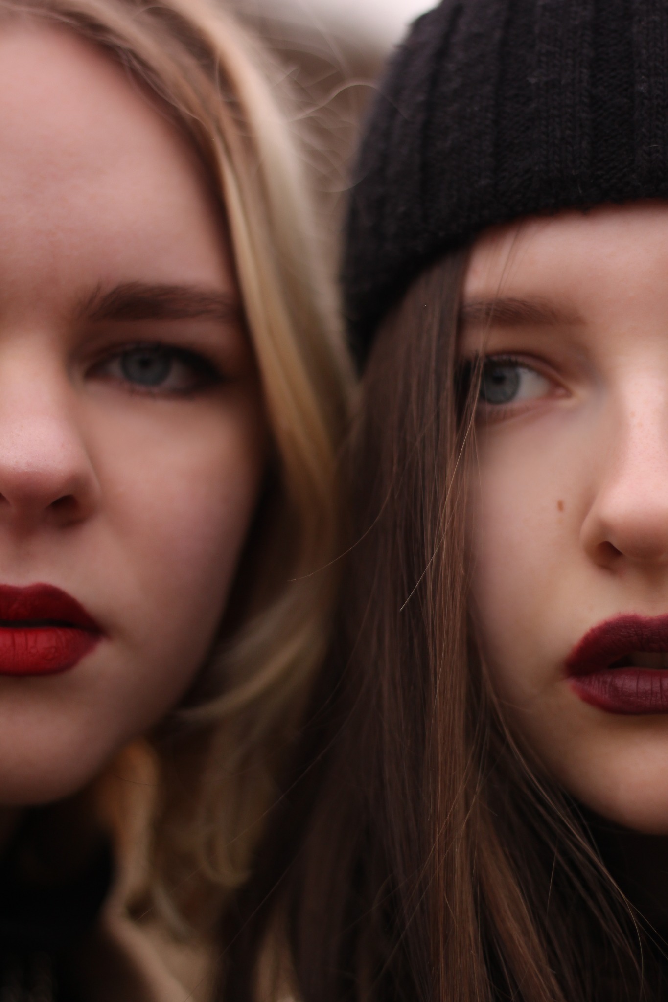 Portret dwóch młodych kobiet, zbliżenie na twarze z mocnym makijażem ust, jedna z blond włosami, druga w czarnej czapce.