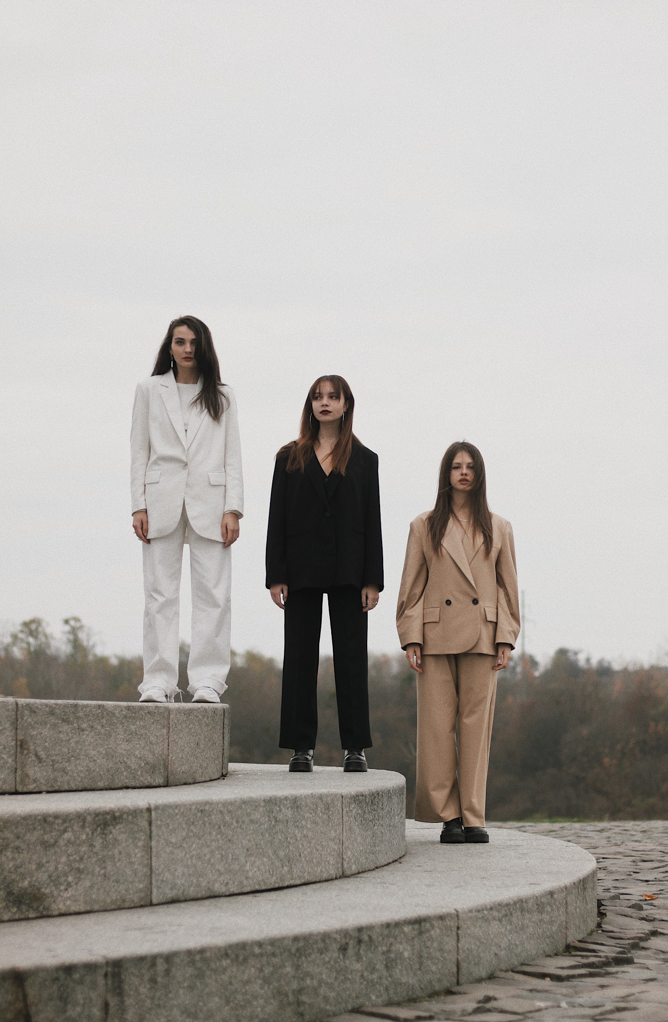 Trzy kobiety w garniturach (białym, czarnym i beżowym) stoją na betonowych schodach na tle pochmurnego nieba i drzew.