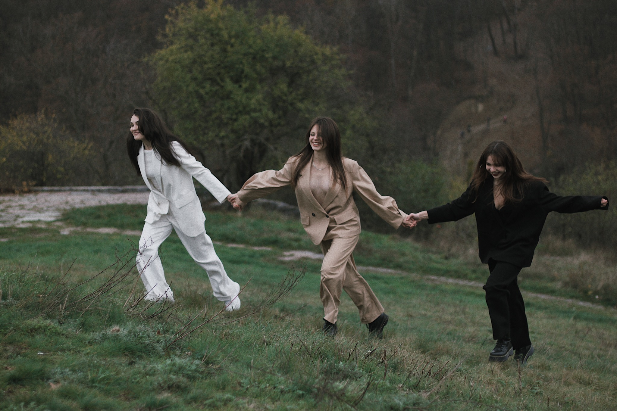 Trzy uśmiechnięte kobiety w garniturach (białym, beżowym i czarnym) biegną trzymając się za ręce po zielonym wzgórzu, jesienne tło z drzewami.