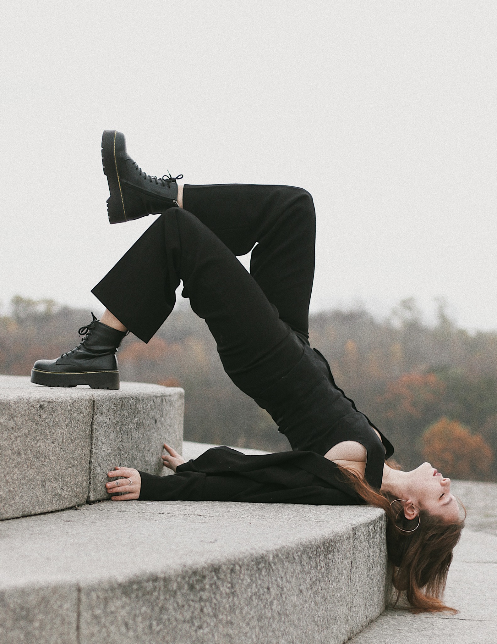 Sesja w Warszawie: Kobieta w czarnym kombinezonie i botkach na platformie, leżąca na kamiennych stopniach, wygina się w łuk, z jesiennym drzewostanem w tle.