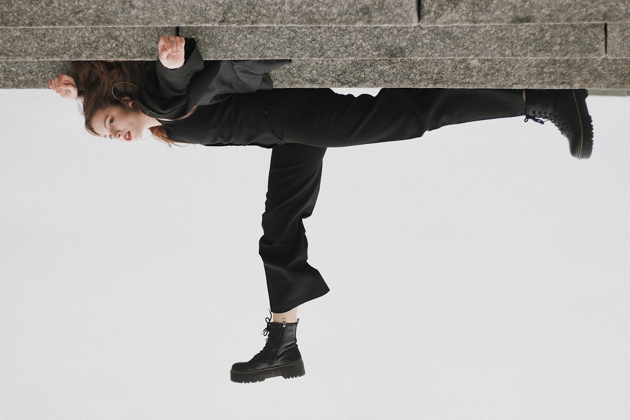 Młoda kobieta w czarnym stroju pozuje na tle jasnego nieba, opierając się o szary, kamienny mur, ujęcie z dołu.