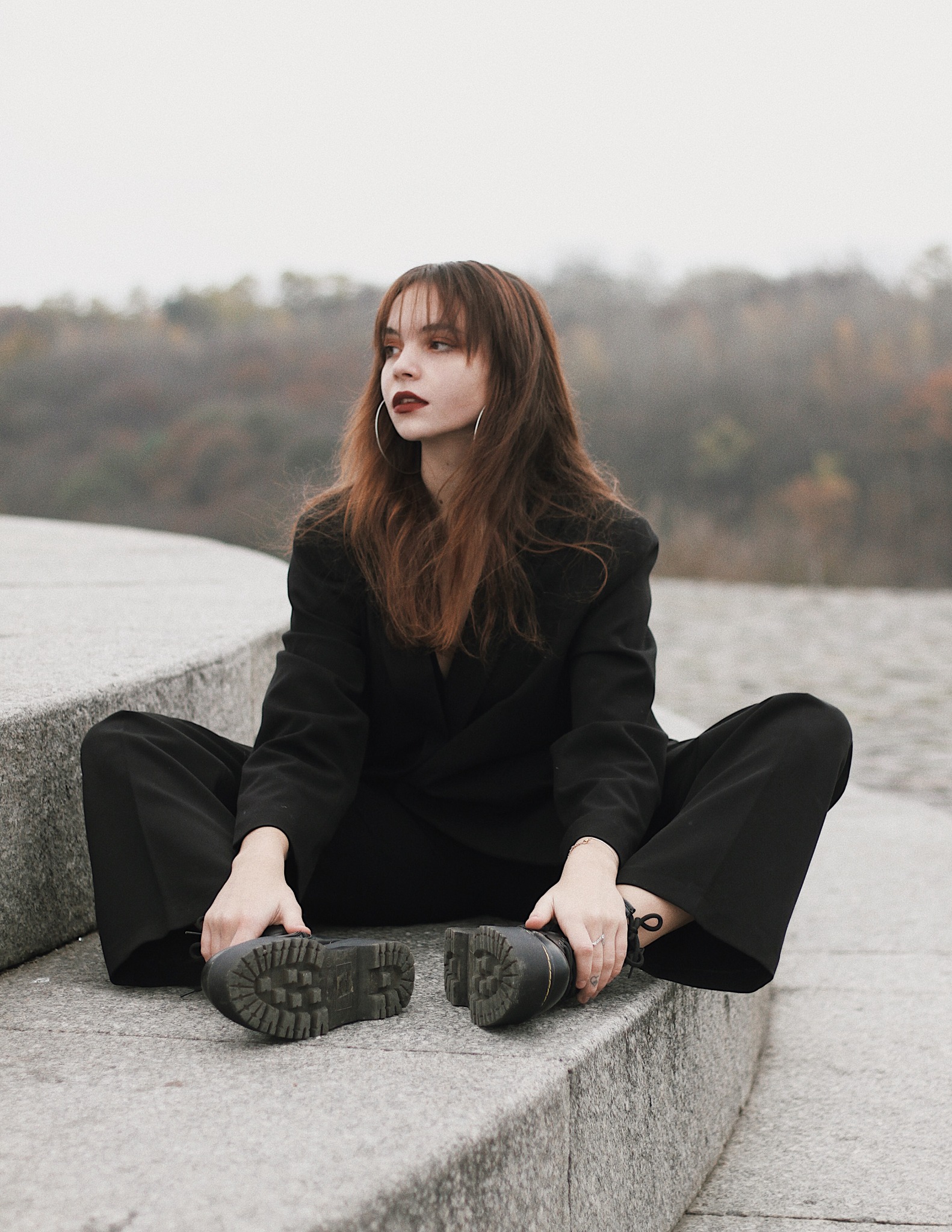 Kobieta w czarnym garniturze i butach typu traper siedzi na betonowych schodach, z zamyślonym wyrazem twarzy, w tle drzewa i mgliste niebo.