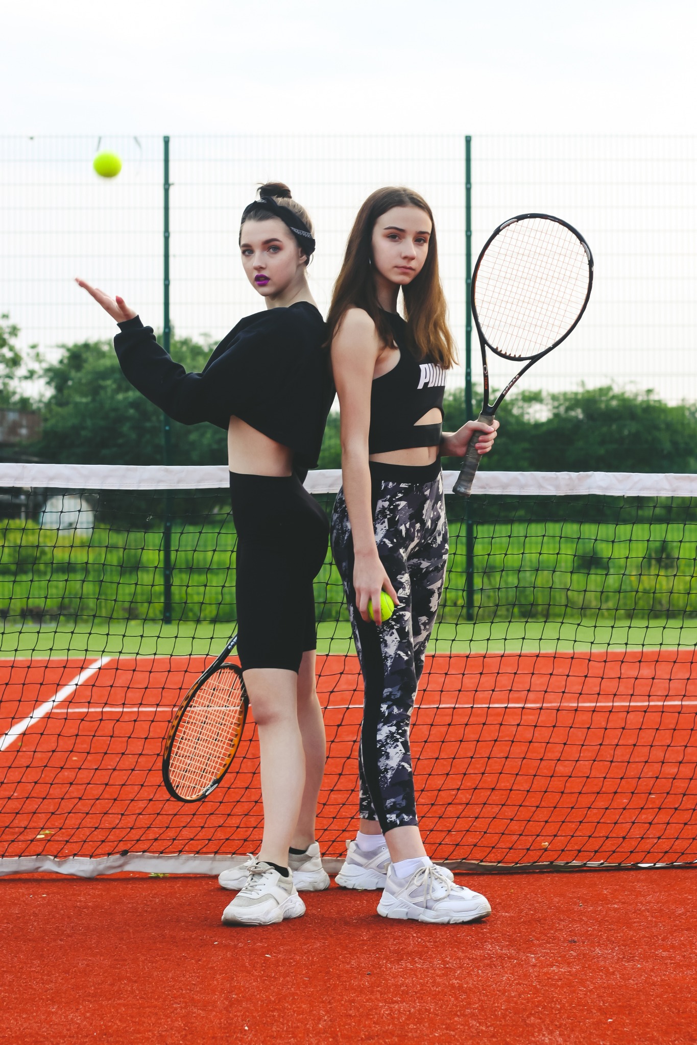 Dwie młode kobiety, ubrane w sportowe stroje, stoją na korcie tenisowym plecami do siebie. Jedna z rakietą i piłką, druga z podniesioną ręką, jakby łapała piłkę w locie. Intensywne oświetlenie...