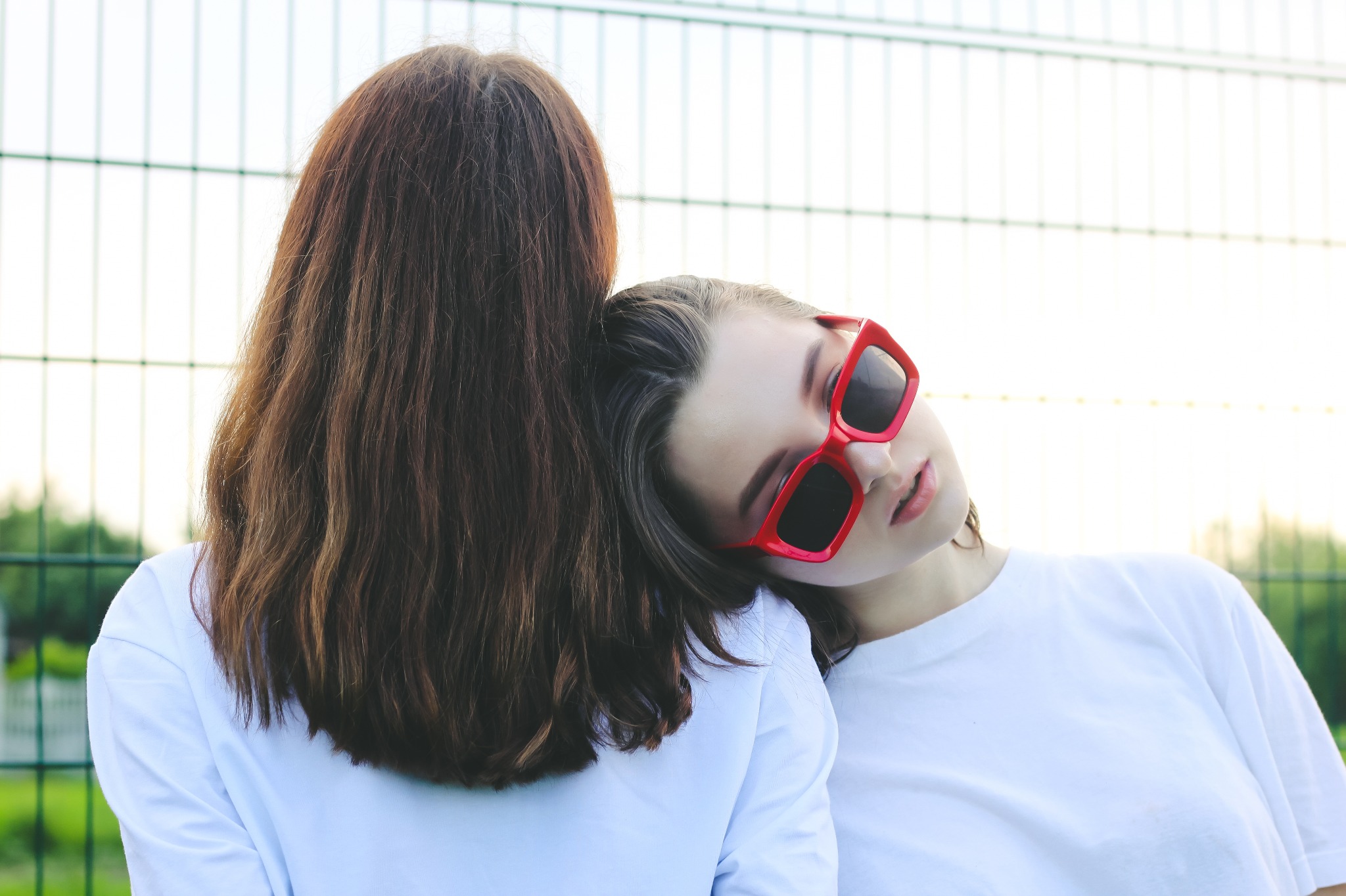 Dwie kobiety w białych koszulkach, jedna opiera głowę na ramieniu drugiej, nosi czerwone okulary, tło stanowi metalowa siatka.