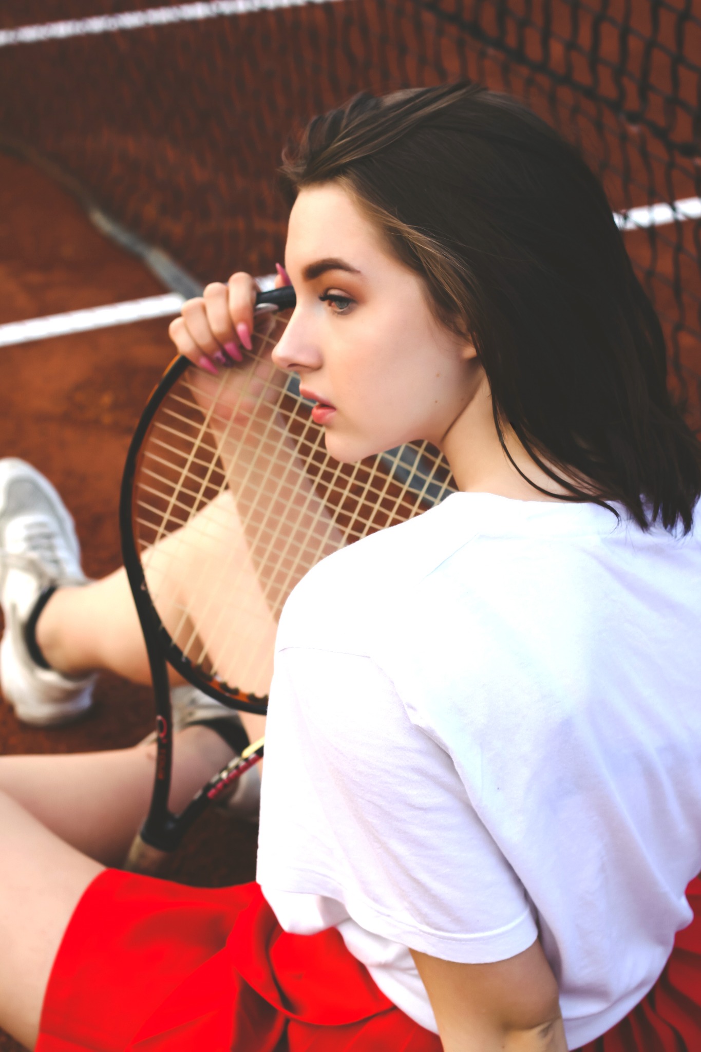 Portret młodej kobiety z rakietą tenisową na korcie, profil, biała koszulka, czerwona spódniczka, sportowy look.