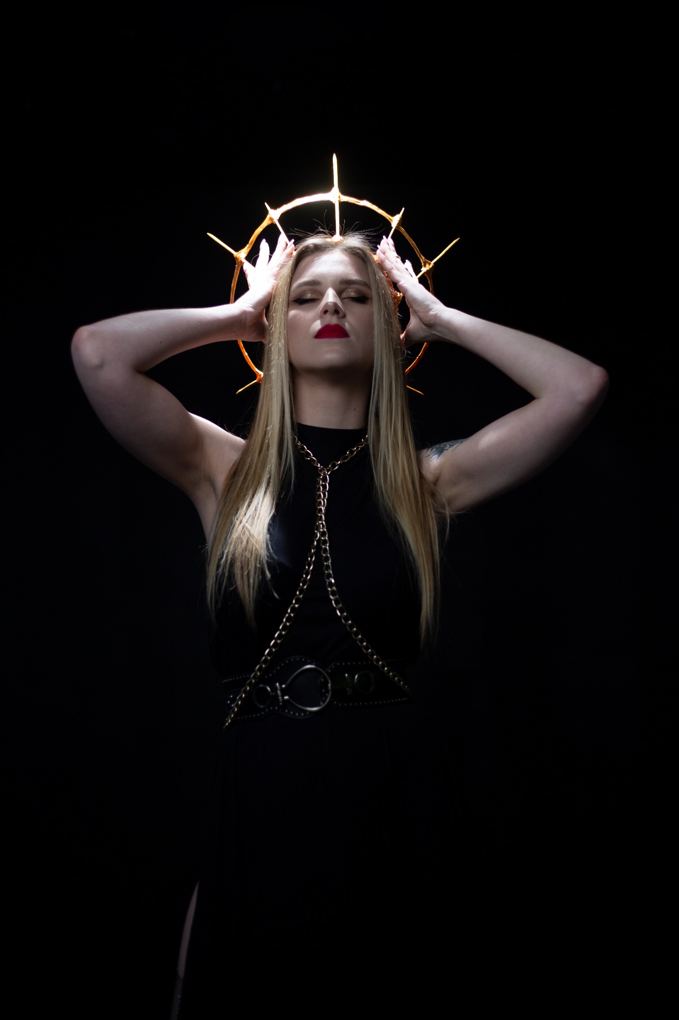 Kobieta z blond włosami, w czarnej sukience z łańcuchem i kolczastą aureolą nad głową, pozuje na ciemnym tle, ręce uniesione do góry.