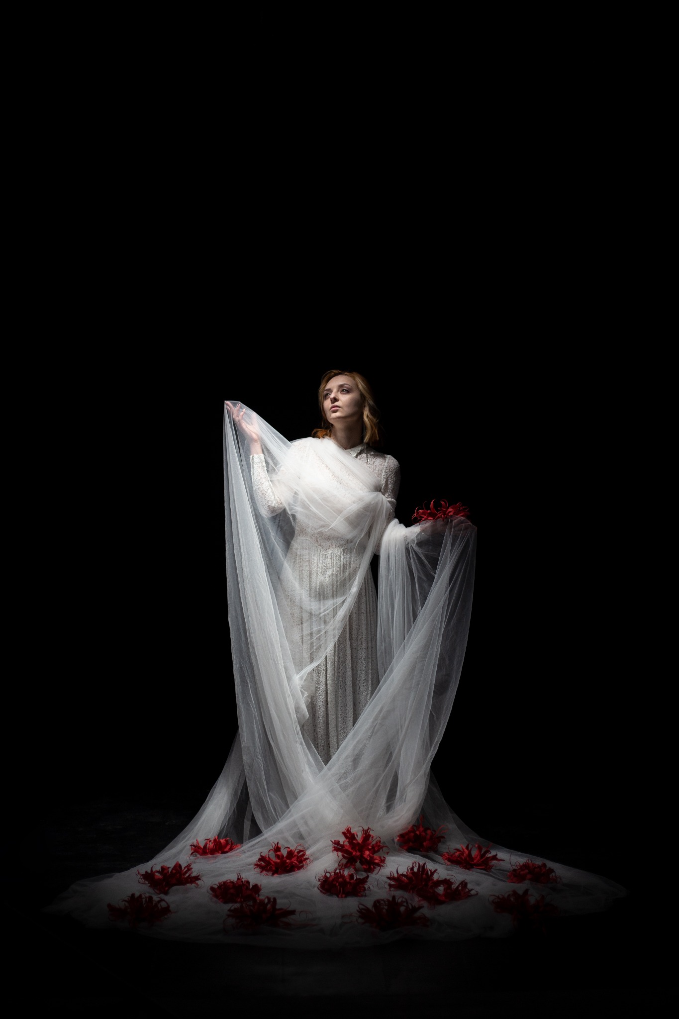 Kobieta w białej sukni z welonem otoczona czerwonymi kwiatami na czarnym tle, patrzy w górę.