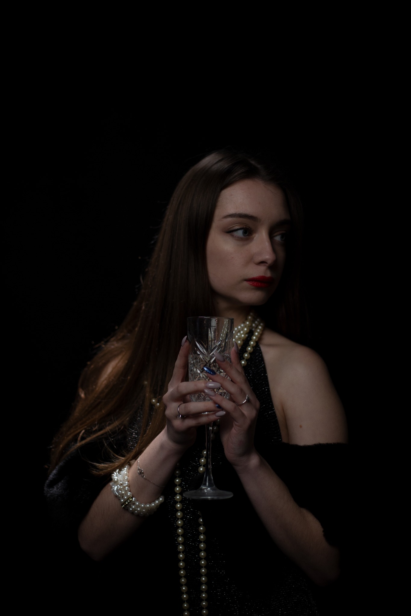 Portret kobiety z długimi brązowymi włosami, w perłach, trzymającej kryształowy kieliszek na ciemnym tle, z delikatnym makijażem i czerwonymi ustami, spoglądającej w bok.