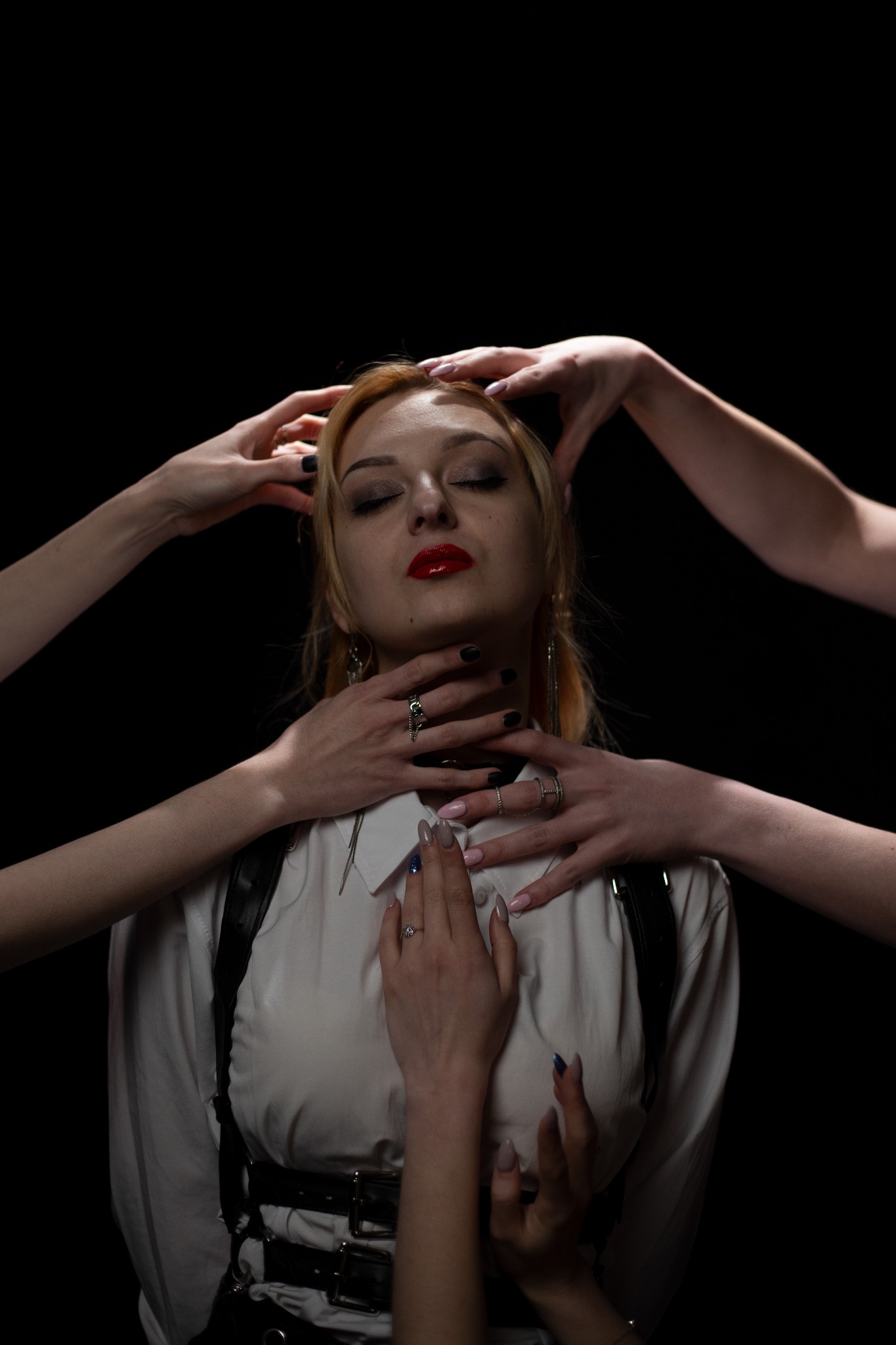 Portret kobiety z zamkniętymi oczami, otoczonej dłońmi w eleganckiej stylizacji z mocnym makijażem na czarnym tle, z widoczną biżuterią na dłoniach.