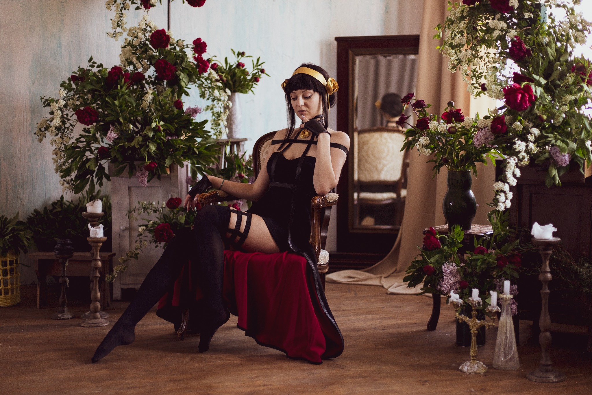 Kobieta w czarnej sukience i złotej opasce siedzi na krześle otoczona kompozycjami kwiatowymi z czerwonych piwonii i białych drobnych kwiatów, w tle lustro i antyczne meble.