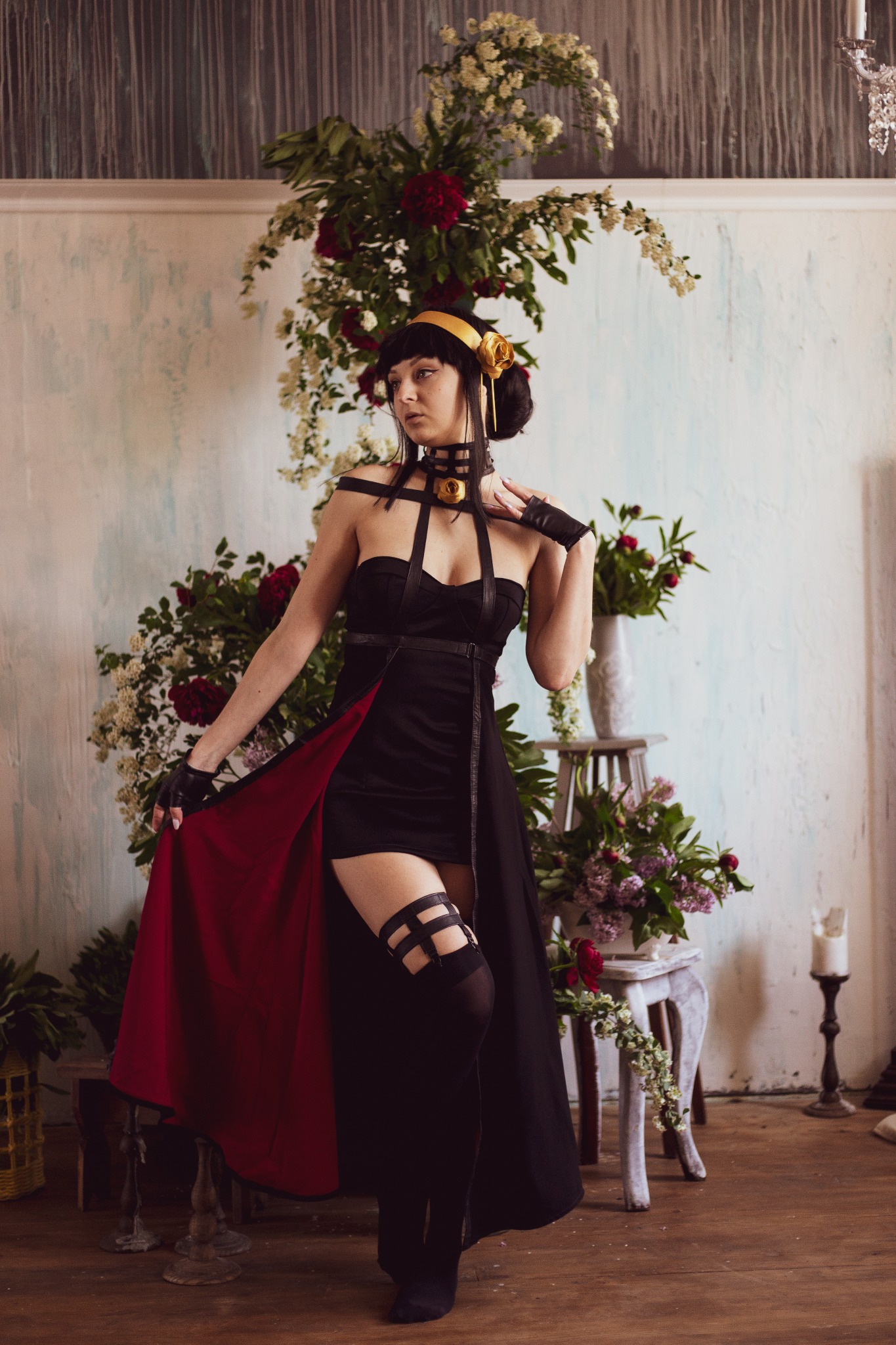 Kobieta w czarnej sukience z czerwonym podszyciem, pozująca na tle dekoracji kwiatowych i stylizowanego wnętrza.