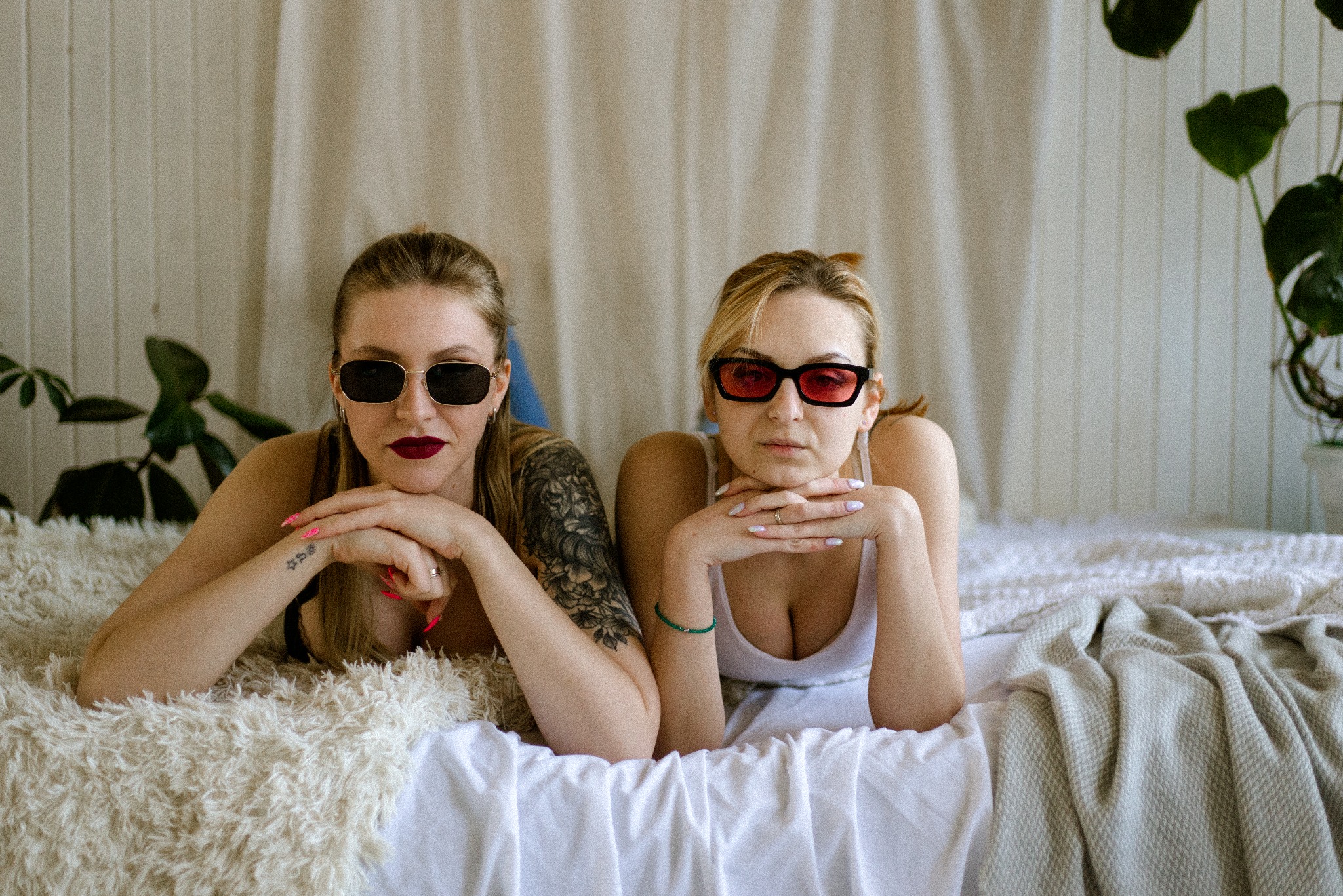 Dwie kobiety w okularach przeciwsłonecznych leżą na łóżku z futrzaną narzutą, pozując i opierając brody na dłoniach, z roślinami w tle.