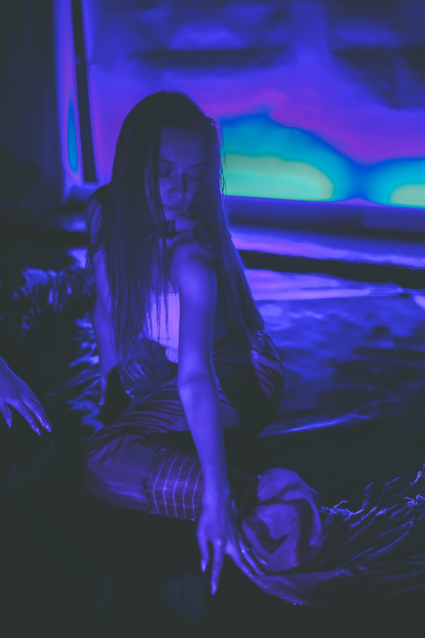 Kobieta siedząca w wodzie w fioletowym oświetleniu, z ręką wyciągniętą w stronę wody, w tle abstrakcyjna grafika.