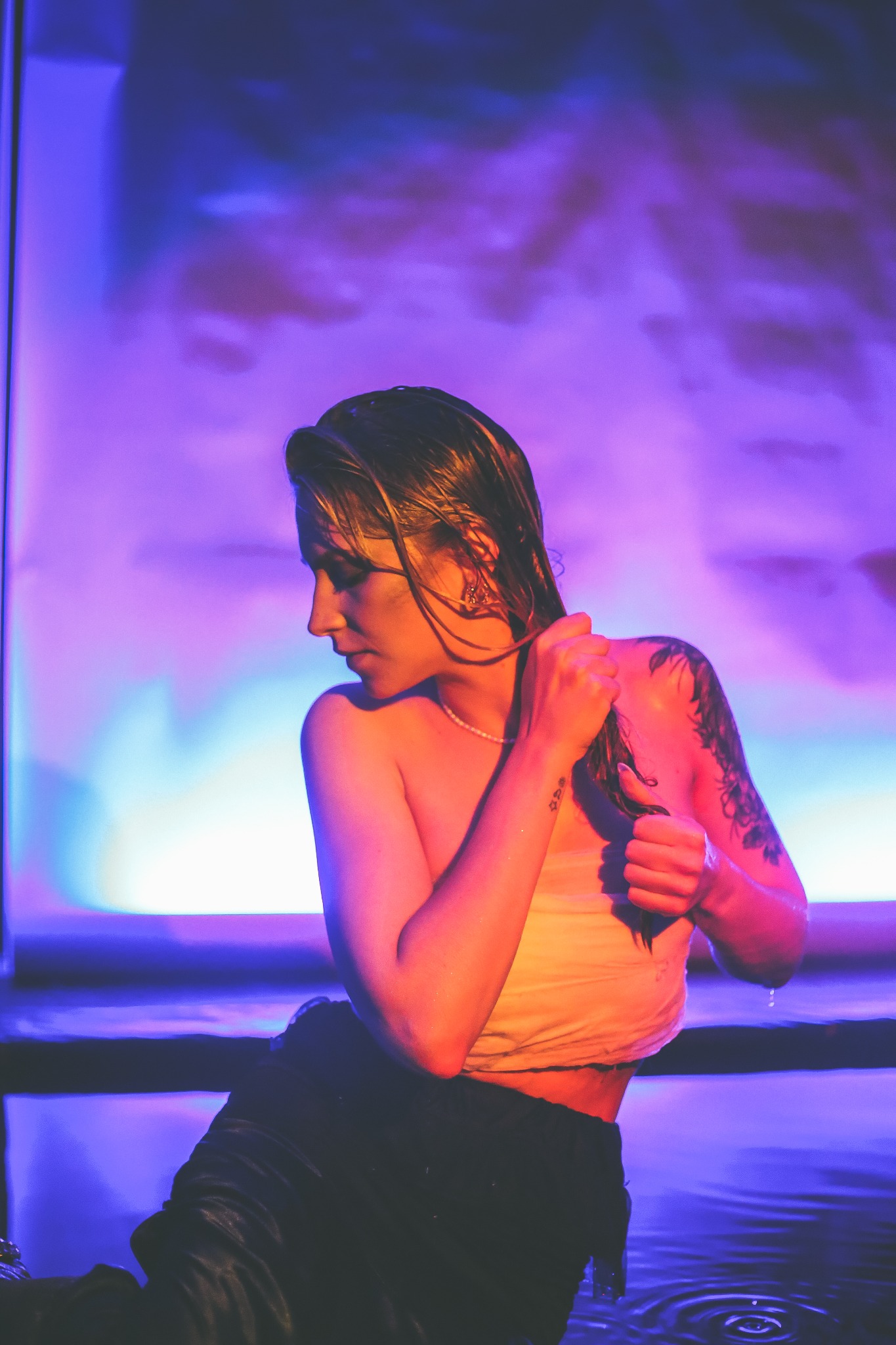 Kobieta z mokrymi włosami, w wodzie, z tatuażami na ramieniu i przedramieniu, w profilu, oświetlona fioletowym światłem.