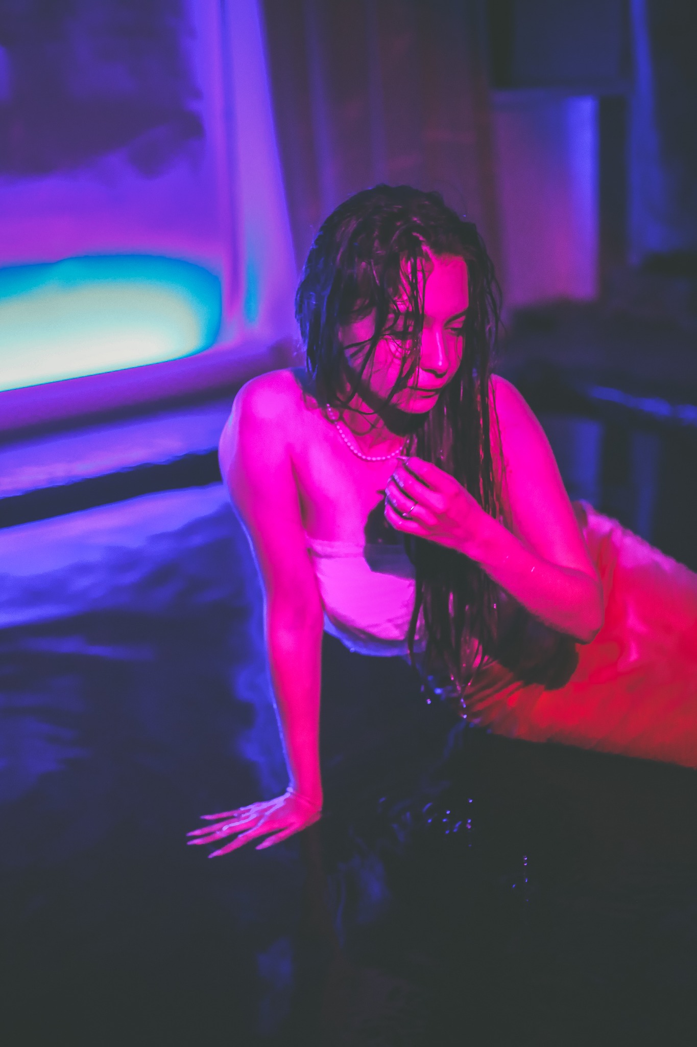 Mokra kobieta w wodzie, oświetlona neonowym różem i fioletem, z perłowym naszyjnikiem, delikatnie dotyka włosów