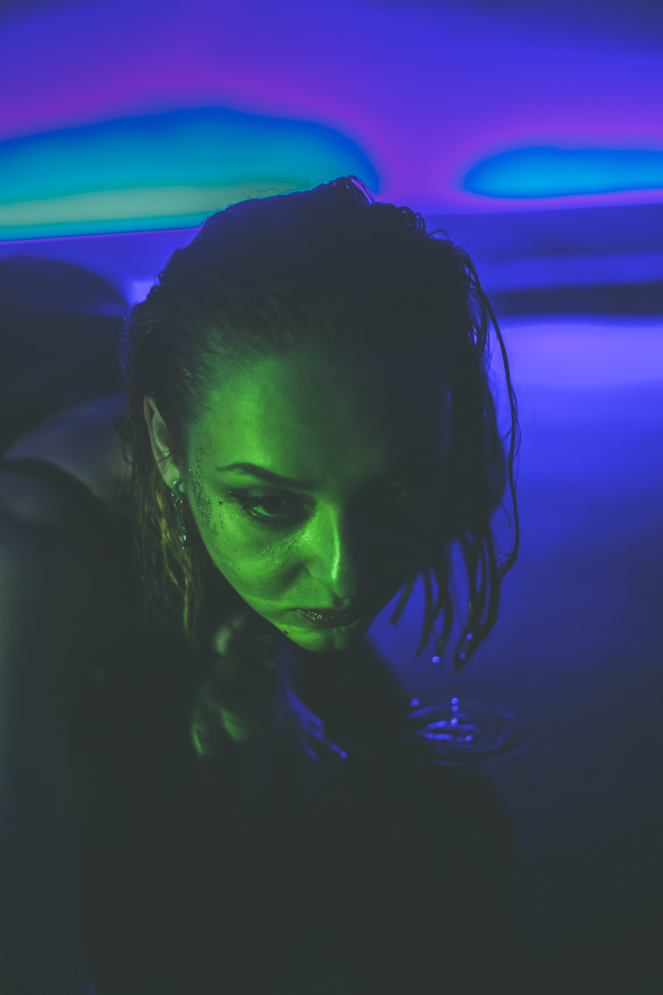 Portret kobiety z mokrymi włosami, zanurzonej w wodzie, oświetlonej zielonym i niebieskim światłem, z kroplami wody spływającymi z rąk.