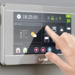 Dotykowy panel sterowania systemem zabezpieczeń marki Satel, zamontowany na ścianie, z widocznym interfejsem użytkownika i dłonią wskazującą na ikonę rolet, sugerującą sterowanie automatyką domową.