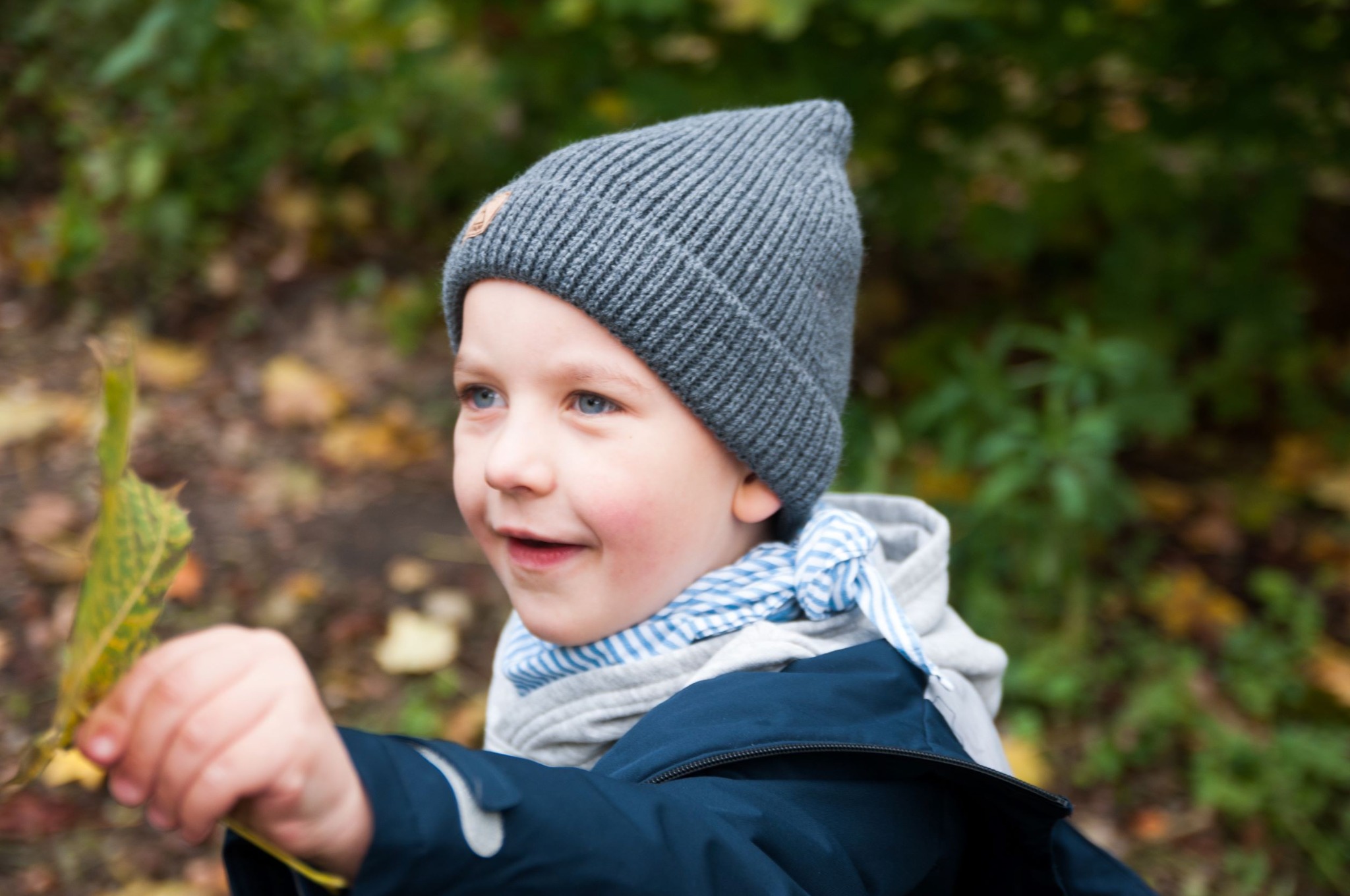 Uśmiechnięty chłopiec w szarej czapce z małą naszywką, w niebieskiej kurtce, trzymający żółty liść na tle zieleni.