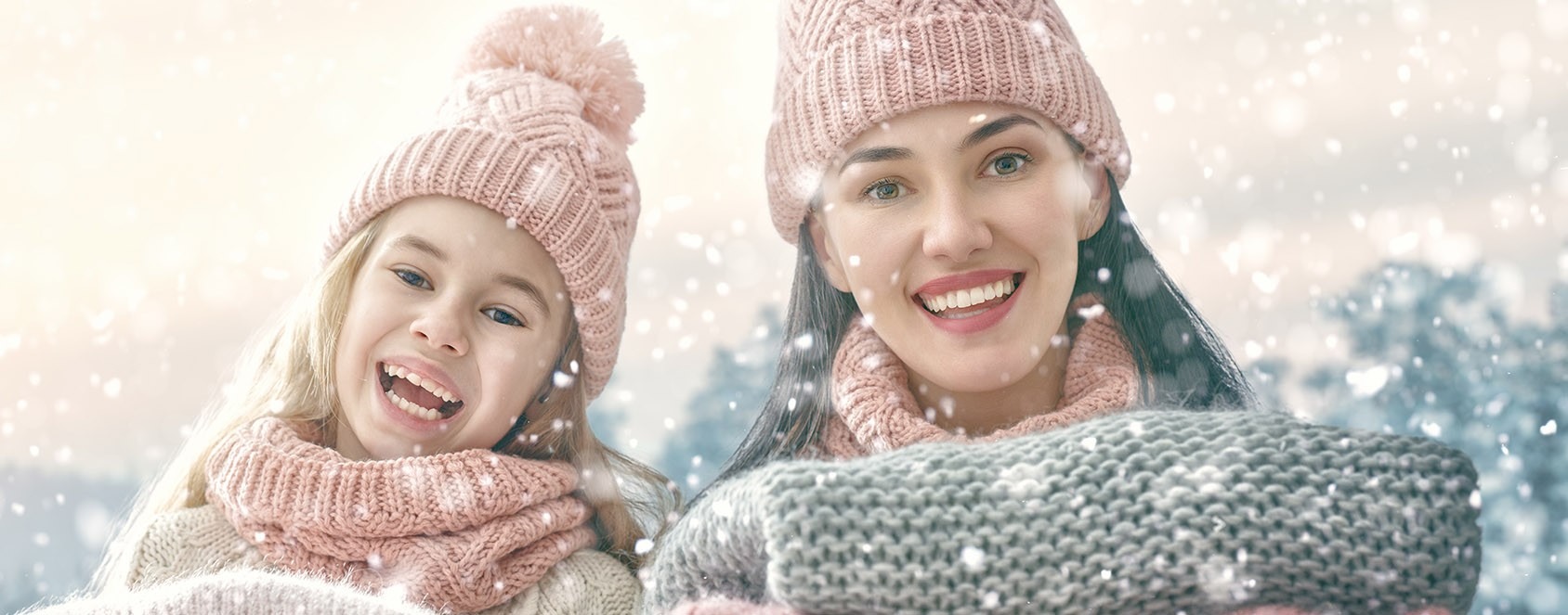 Uśmiechnięta matka i córka w różowych czapkach i szalikach, śnieg pada, zimowy krajobraz w tle.