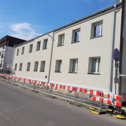 DYSTRYBUCJA 88 Sp. z o.o. - Świeżo otynkowany budynek wielorodzinny z jasną elewacją, zabezpieczony barierkami z czerwono-białymi oznaczeniami, widoczny fragment rusztowania i znaki drogowe.