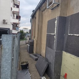 DYSTRYBUCJA 88 Sp. z o.o. - Układanie grafitowych płyt izolacyjnych na ścianie budynku, widoczna piana montażowa, wiadro z zaprawą i narzędzia murarskie, fragment metalowej konstrukcji przy ścianie.