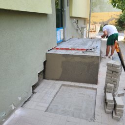 DYSTRYBUCJA 88 Sp. z o.o. - Prace murarskie przy wejściu do budynku: wyrównywanie powierzchni betonowej schodów i układanie kostki brukowej na chodniku wejściowym, widoczny poziomica, kielnia i zapas kostki.