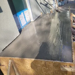 Świeżo wylana, mokra betonowa wylewka w drewnianym szalunku, przy budynku z ocieploną elewacją zabezpieczoną kartonami, widoczne narzędzia i kontener na gruz w tle.