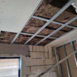 DYSTRYBUCJA 88 Sp. z o.o. - Montaż ocieplenia poddasza z wełny mineralnej i metalowego stelaża, częściowo pokrytego płytą gipsową, widok na surową ścianę z pustaków i zabezpieczone okno.