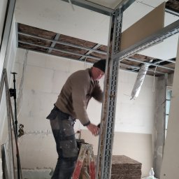 DYSTRYBUCJA 88 Sp. z o.o. - Pracownik na drabinie montuje metalowy stelaż sufitu podwieszanego, widoczne płyty gipsowo-kartonowe i izolacja z wełny mineralnej.