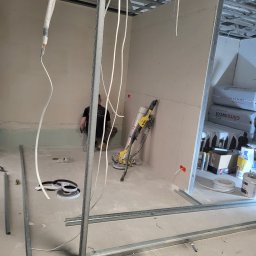 DYSTRYBUCJA 88 Sp. z o.o. - Wnętrze w trakcie budowy, widoczne stelaże metalowe, płyty gipsowo-kartonowe, szlifierka do gipsu oraz pracownik przy ścianie.