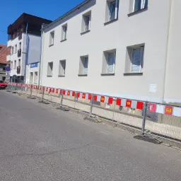 Biała elewacja budynku po remoncie, zabezpieczona barierkami ochronnymi na ulicy, widoczne okna i fragment sąsiedniego budynku.