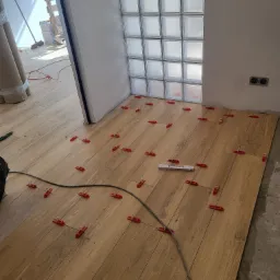 Układanie drewnopodobnych płytek podłogowych z użyciem czerwonych klipsów poziomujących, widoczna ściana z luksferami i niezakończona podłoga.